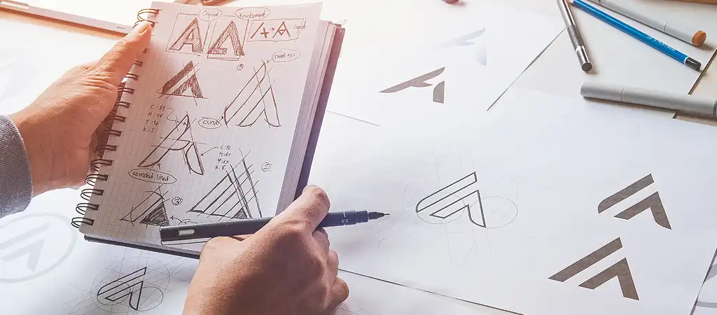 A logo grafikai tervezésének folyamata, papírra lerajzolva több féle vázlat és terv színkódokkal és arányokkal együtt.
